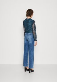 Tyrkysová krajková halenka s dlouhými rukávy, vroubkovaným výstřihem a klíčovým otvorem na zádech, v kombinaci s vysokým pasem, světle modrými džínami s širokými nohavicemi a černými podpatky.