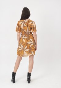 Mujer con vestido de manga corta hasta la rodilla con grandes patrones florales en naranja y beige, combinando con botines negros de tacón, de espaldas.