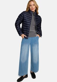Doudoune marine avec coutures horizontales, haut rayé, large pantalon en denim bleu clair et mocassins noirs, portée par une personne debout.