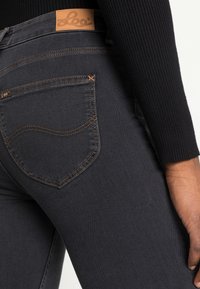 Jeans de mezclilla negra con un diseño ajustado, que presentan un parche de cuero marrón y costuras marrones en contraste en los bolsillos traseros.