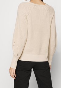 Beżowy sweter dziany z prążkowanymi mankietami i dołem, z teksturowanym wzorem i otwartymi detalami na rękawach. Noszony z czarnymi spodniami.