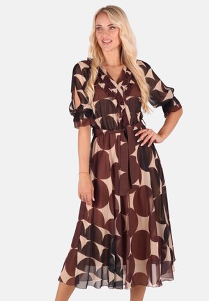 Langes, transparentes Kleid mit einer Mischung aus großen braunen und beigen Kreisen, Rüschenausschnitt und einem gebundenen Taillengürtel. Die Ärmel sind gepufft.