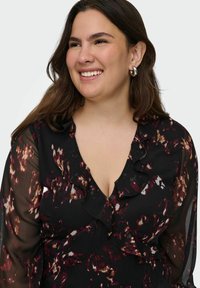 Mujer sonriente con cabello oscuro que lleva pendientes de aro plateados y una blusa negra transparente con un patrón floral en tonos burdeos y blanco, y cuello con volantes.