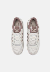 Balti un gaiši bež sporta apavi ar trim brūnām līnijām, caurumiņiem pie pirkstiem un mīkstu apkaklīti. Iekļauj auklu aizdari un apzīmējuma logo.