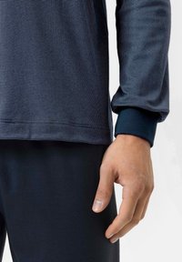 Granatowa koszula z długim rękawem o teksturowanym wzorze, z eleganckim wykończeniem i dopasowanymi mankietami, w połączeniu z ciemnymi, pasującymi spodniami oraz swobodną pozycją rąk.