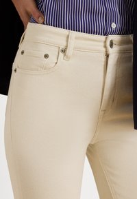 Calças de ganga bege com textura suave, design de cinco bolsos e detalhes em metal. Apresentam fecho de correr e corte ajustado.