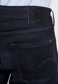 Mörkblå denimjeans med en slät yta, med en svart läderlapp, bakfickor och subtila sömnadsdetaljer.