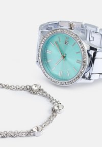 Montre en argent avec un cadran turquoise et des accents en cristal ; à côté, un bracelet en chaîne argenté orné de trois embellissements en pierres précieuses transparentes.