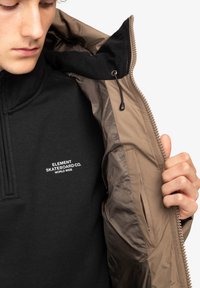 Maglione nero con colletto zip caratterizzato da un marchio bianco. Sopra di esso è indossata una giacca marrone imbottita con una texture liscia e dettagli con zip.
