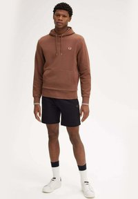 Felpa in cotone marrone con una tasca frontale, abbinata a pantaloni corti neri. Presenta polsini a strisce bianche e sneakers bianche. Logo semplice sulla felpa.