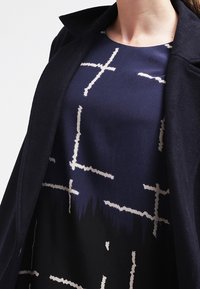 Laiva tunika, jossa on abstrakti kerma kuvio leikkaavista viivoista, kerrostettuna tumman kankaan takin alle, esittelee sileää tekstuuria ja rakenteellista muotoilua.