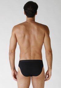 Modello maschile in piedi di spalle, indossando un perizoma nero, mostrando la schiena nuda, braccia rilassate ai lati su uno sfondo chiaro e semplice.