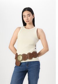 Brun cuirbelte med sirkulære detaljer og et sentralt dekorativt element. Modellen har på seg en lys ribbestrikket tank-top og blå jeans.