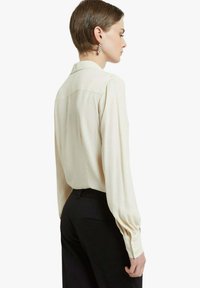 Camicia a maniche lunghe color crema con una texture morbida, polsini con bottoni e cucitura sulla spalla posteriore, abbinata a pantaloni neri.