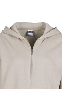Lys sandfarget zip-hettegenser med snorhette, myk tekstur, og et stoffmerke som står "URBAN CLASSICS" inni halsen.