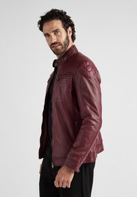 Burgunderfarbene Lederjacke mit gesteppte Schulterakzenten, vorderen Reißverschlusstaschen und Stehkragen; strukturierte Oberfläche und enganliegendes Design.