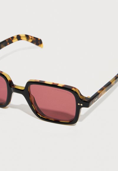 Γυαλιά ηλίου με σχέδιο χελώνας, ορθογώνιους φακούς με κόκκινη απόχρωση και λεπτούς βραχίονες, τοποθετημένα σε λευκή επιφάνεια.