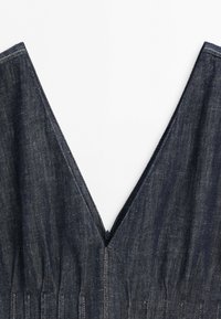 Gros plan d'un tissu en denim foncé avec un décolleté en V profond et des coutures visibles sur fond blanc.