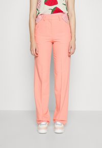 Vrouw draagt koraalkleurige, recht gesneden broek met hoge taille, gecombineerd met pastelkleurige sneakers en een mouwloos topje met aardbeiendessin.