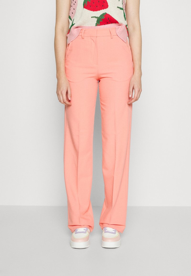 Vrouw draagt koraalkleurige, recht gesneden broek met hoge taille, gecombineerd met pastelkleurige sneakers en een mouwloos topje met aardbeiendessin.