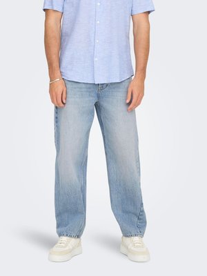 Mann trägt hellblaues kurzärmeliges Hemd mit Knöpfen, hell gewaschene, locker sitzende Jeans, weiße Sneakers und ein silbernes Armband am linken Handgelenk.