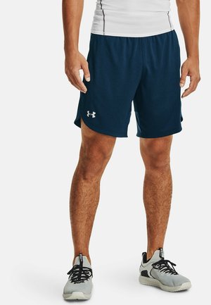 Mężczyzna noszący granatowe sportowe spodenki z logo Under Armour, białą koszulkę kompresyjną oraz szare buty do biegania z czarnymi sznurówkami na białym tle.