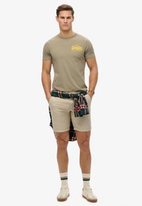 Tan kortærmet t-shirt med gul tekst, beige shorts og en grøn/rød ternet skjorte bundet om taljen; creme sneakers og stribede sokker.