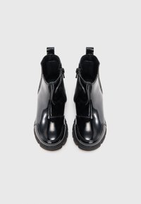 Ζεύγος μαύρων γυαλιστερών δερμάτινων μποτών μέχρι τον αστράγαλο με χοντρές σόλες και πλευρικούς φερμουάρ, εμφανιζόμενες από πάνω σε λευκό φόντο.