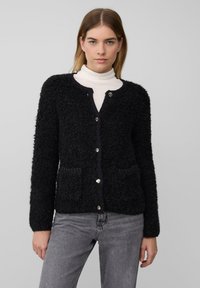 Schwarze strukturierte Strickjacke mit langen Ärmeln, rundem Ausschnitt, zwei Taschen vorne und silbernen Knöpfen. Darüber wird ein weißer Rollkragenpullover getragen.