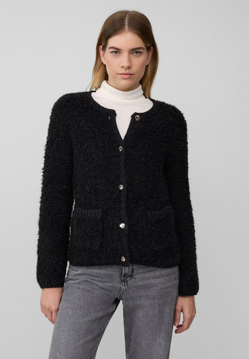 Schwarze strukturierte Strickjacke mit langen Ärmeln, rundem Ausschnitt, zwei Taschen vorne und silbernen Knöpfen. Darüber wird ein weißer Rollkragenpullover getragen.