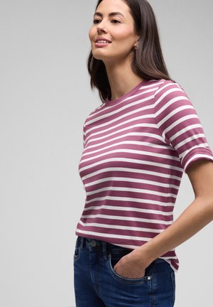 Mujer sonriendo, vistiendo una camiseta de manga corta a rayas moradas y blancas y jeans azules, con una mano en el bolsillo, sobre un fondo liso.