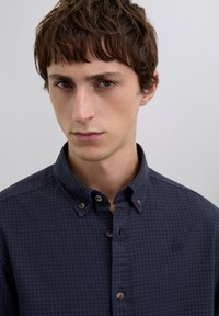 Camisa azul marino a cuadros con cuello clásico, cierre delantero de botones y un detalle de logo sutil. La tela parece suave con un acabado ligeramente texturizado.