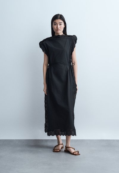 Γυναίκα που στέκεται σε γκρι πάτωμα μπροστά σε ανοιχτόχρωμο τοίχο, φορώντας μαύρο μεσαίο φόρεμα με κυματιστά δαντελένια τελειώματα και καφέ φλατ σανδάλια.