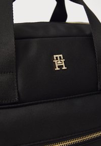 Sac en tissu noir avec un logo doré, des détails de fermeture éclair et des poignées solides. La surface a une apparence texturée et tissée.