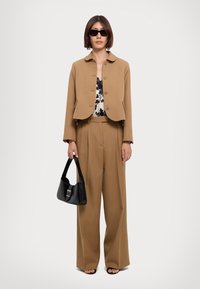 Brown cropped jacket over a floral patterned blouse, wide-leg brown pants, and black handbag. Model also wears black sunglasses and sandals.