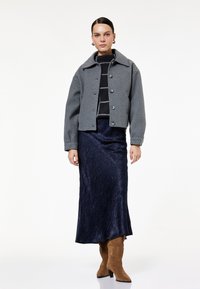 Grå uldjakke med stor krave og knapper, parret med en navy satin nederdel, knæhøje lysebrune ruskindsstøvler og en stribet striktrøje.