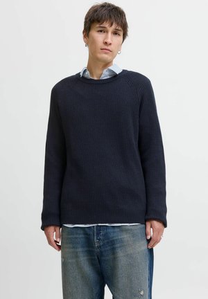 Pull en tricot côtelé bleu marine avec des manches longues, un col rond et une coupe décontractée, superposé sur une chemise rayée bleu et blanc.