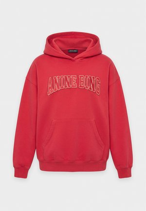 Femme portant un sweat à capuche rouge et un pantalon, tenant un sac fourre-tout à motif zèbre noir et blanc, debout devant un fond blanc uni.