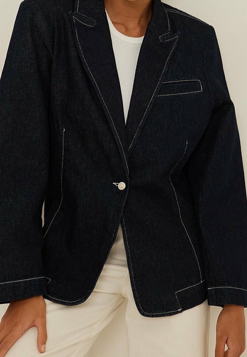 Personne portant un blazer en denim foncé avec des surpiqûres blanches sur un haut blanc et un pantalon couleur crème, main reposant sur la cuisse.