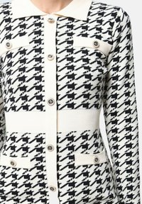 Cardigan à manches longues en motif pied-de-poule noir et blanc avec col crème, boutons décoratifs et rabats de poche.