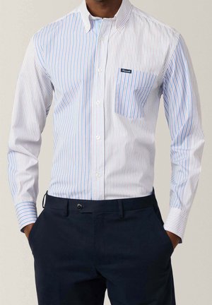 Mand iført hvid langærmet skjorte med lodrette blå og røde striber og marineblå bukser, hænderne i lommerne, står mod en ensfarvet baggrund.