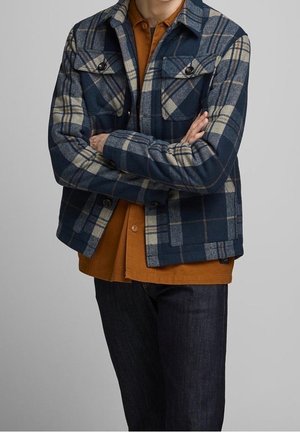 Veste à carreaux en mélange de laine bleu marine et beige, avec deux poches poitrines, boutons noirs et coupe décontractée sur une chemise orange. Jean foncé porté en dessous.