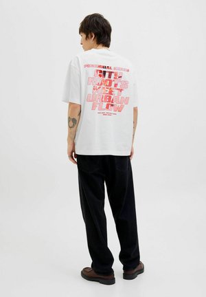 Hvid t-shirt med korte ærmer, der har stor rød grafisk tekst på ryggen. Sæt sammen med sorte bukser og brune sko.