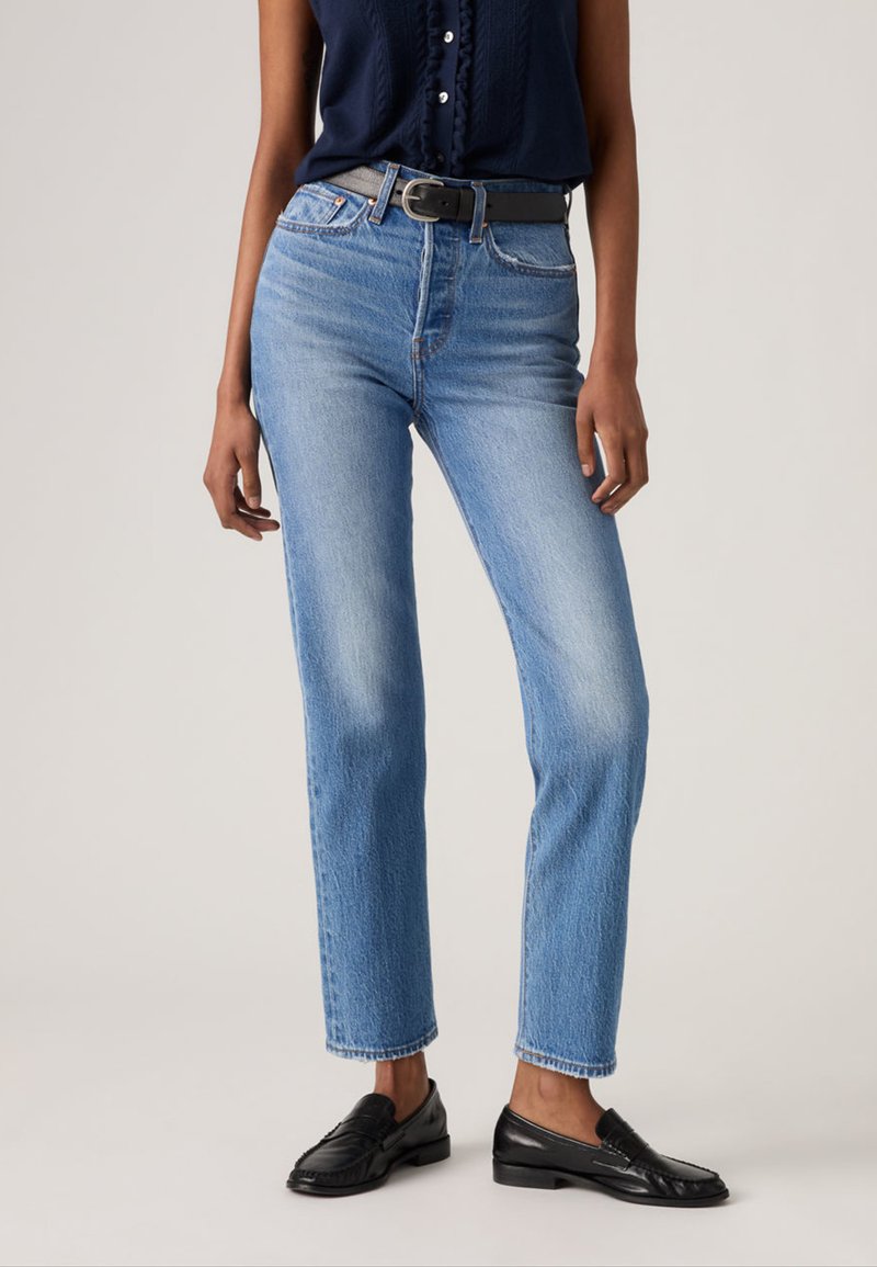 Γυναίκα που φοράει ανοιχτόχρωμα τζιν ψηλής μέσης με μαύρη ζώνη, μπλε σκούρο αμάνικο τοπ και μαύρα δερμάτινα μοκασίνια σε απλό φόντο.