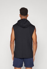 Schwarze ärmellose Kapuzenjacke mit weicher Textur, ausgestattet mit einer geräumigen Kapuze, geschichtetem Schulterdesign und lockerem Schnitt. Trägt dazu navyfarbene Shorts.