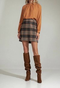Blusa a maniche lunghe color ruggine con scollatura arricciata abbinata a una mini gonna a quadri nei toni marrone e beige. Stivali alti fino al ginocchio in suede marroni.