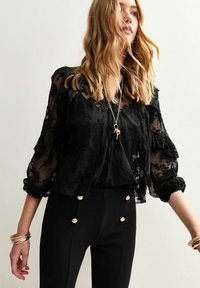 Femme portant un chemisier en dentelle noire transparente avec des manches bouffantes, un pantalon noir avec des boutons dorés, des bracelets dorés et un collier avec un pendentif long.