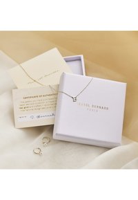 Gold necklace with a "B" pendant, placed inside a light purple box. Also features a certificate of authenticity and pearl-accented hoop earrings.