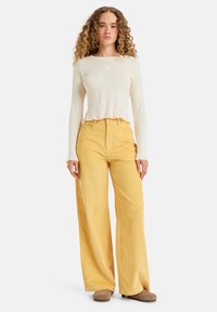 Un haut beige clair à manches longues avec une texture côtelée et un ourlet en forme de vagues, associé à un pantalon large en velours côtelé jaune taille haute. Chaussures marron.