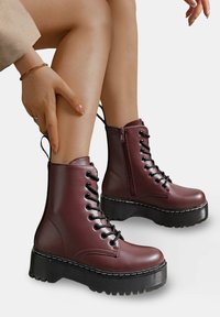 Bottines en simili cuir bordeaux avec lacets noirs, une fermeture éclair sur le côté et de grosses semelles noires avec un motif de crampons texturé.